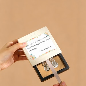 May include: A square sliding jewelry box with a cream-colored outer sleeve featuring a floral border and the text: You can customize your own blessings or personal message Your Name. Inside, a black frame holds two clear teardrop earrings.
