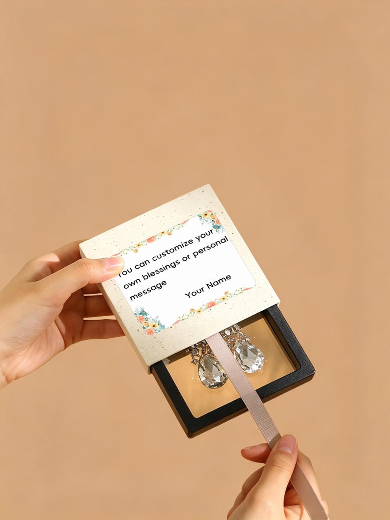 May include: A square sliding jewelry box with a cream-colored outer sleeve featuring a floral border and the text: You can customize your own blessings or personal message Your Name. Inside, a black frame holds two clear teardrop earrings.