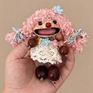 May include: A small doll with curly pink hair, a red nose, and a wide open mouth. The doll wears a white lace dress adorned with small blue and pink crocheted flowers. Its arms and legs are made of dark brown beads, and light blue bows decorate its hair.
