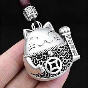 Puede incluir: Un colgante plateado con forma de gato de la suerte, con patrones de filigrana, un motivo de moneda central y un pergamino con caracteres chinos en una pata. Cuelga de un cordón negro con una cuenta de madera oscura y un separador plateado.