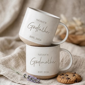 May include: Two off-white ceramic mugs stacked vertically. The top mug features the engraved text "NOAH'S Godfather EST. 2026" and the bottom mug features the engraved text "NOAH'S Godmother EST. 2026". Both mugs have a smooth finish and a light brown base.
