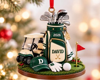 May include: A Christmas ornament featuring a green and cream golf bag with the name DAVID and the year 2026, a golf cart, a green cap with the letter D, a white golf glove, a golf ball, and a red flag on a pole, all on a circular base.
