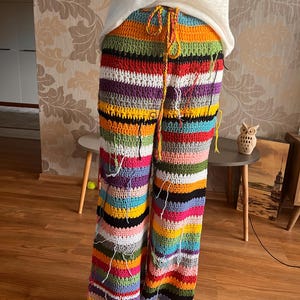 Puede incluir: Pantalones de ganchillo hechos a mano con un vibrante patrón de rayas en varios colores, incluyendo naranja, verde, morado y rosa. Los pantalones tienen una cintura con cordón y un ajuste holgado de pierna ancha. Los extremos del hilo son visibles.