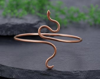 May include: A copper-coloured metal bracelet featuring a continuous wire design shaped like a stylised snake. The bracelet has a smooth, polished finish and a flexible open-ended structure that curves into a serpent form at the centre point.