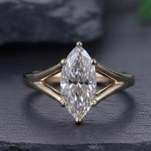 May include: A gold ring featuring a single marquise-cut clear gemstone set in a prong setting. The band has a split shank design that tapers toward the center stone, creating a sleek and symmetrical appearance with a polished metallic finish.