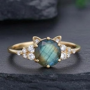 May include: A gold ring featuring a round, iridescent blue-green gemstone, flanked by small white gemstones. The ring's design includes cat ear accents on the gemstone setting. The ring is set against a dark background.