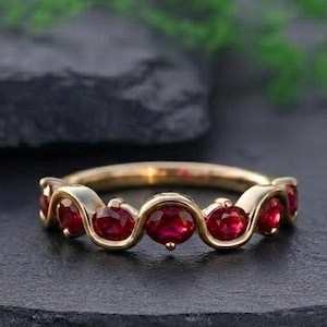 May include: A gold ring featuring a series of oval-cut, vibrant red gemstones. The gemstones are set in a wavy, continuous band, creating a flowing design. The ring is displayed on a dark, textured surface, highlighting its color and design.