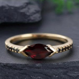 May include: A gold ring with a marquise-cut, deep red gemstone as the centerpiece. The band is adorned with small black stones, and the ring is set against a dark background, showcasing its elegant design.