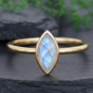 May include: A gold ring featuring a marquise-cut moonstone with iridescent blue and white hues. The gemstone is set in a slender gold band. The ring is presented on a dark grey surface, with green foliage visible in the background.