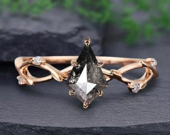 May include: A rose gold ring with a kite-cut salt and pepper diamond. The band has a nature-inspired design with intertwined branches and small, round accent stones. The ring is set against a dark, textured surface.
