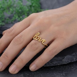 May include: A gold-toned ring featuring the word love written in a continuous cursive script. The metal has a matte finish and the design is formed by a single wire-like band that wraps around the finger to display the word clearly.