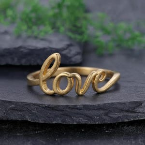 May include: A gold-toned ring featuring the word love written in a continuous cursive script. The metal has a matte finish and the letters are integrated directly into the band, forming the focal point of the jewellery piece.