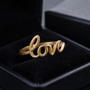 May include: A gold-toned ring featuring the word love written in a continuous cursive script. The ring has a matte, textured finish and is positioned inside a black velvet-lined jewellery box with a matching black exterior lid visible.