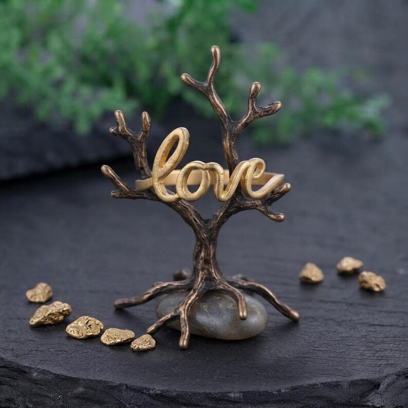 May include: A gold-toned ring featuring the word love in cursive script, displayed on a dark bronze-coloured metal tree-shaped stand. The stand is supported by a smooth grey stone base, surrounded by small, irregular gold-coloured metallic fragments.