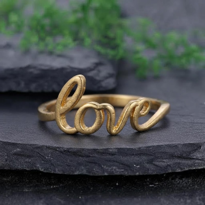 May include: A gold-toned ring featuring the word love written in a continuous cursive script. The metal has a matte finish and the letters are integrated directly into the band, forming the focal point of the jewellery piece.
