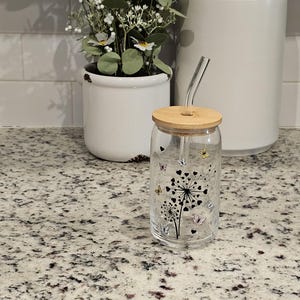 May include: A clear glass tumbler with a bamboo lid and a curved glass straw. The tumbler features a black dandelion design with colorful butterfly accents. A white ceramic pot with greenery and a white canister are in the background.