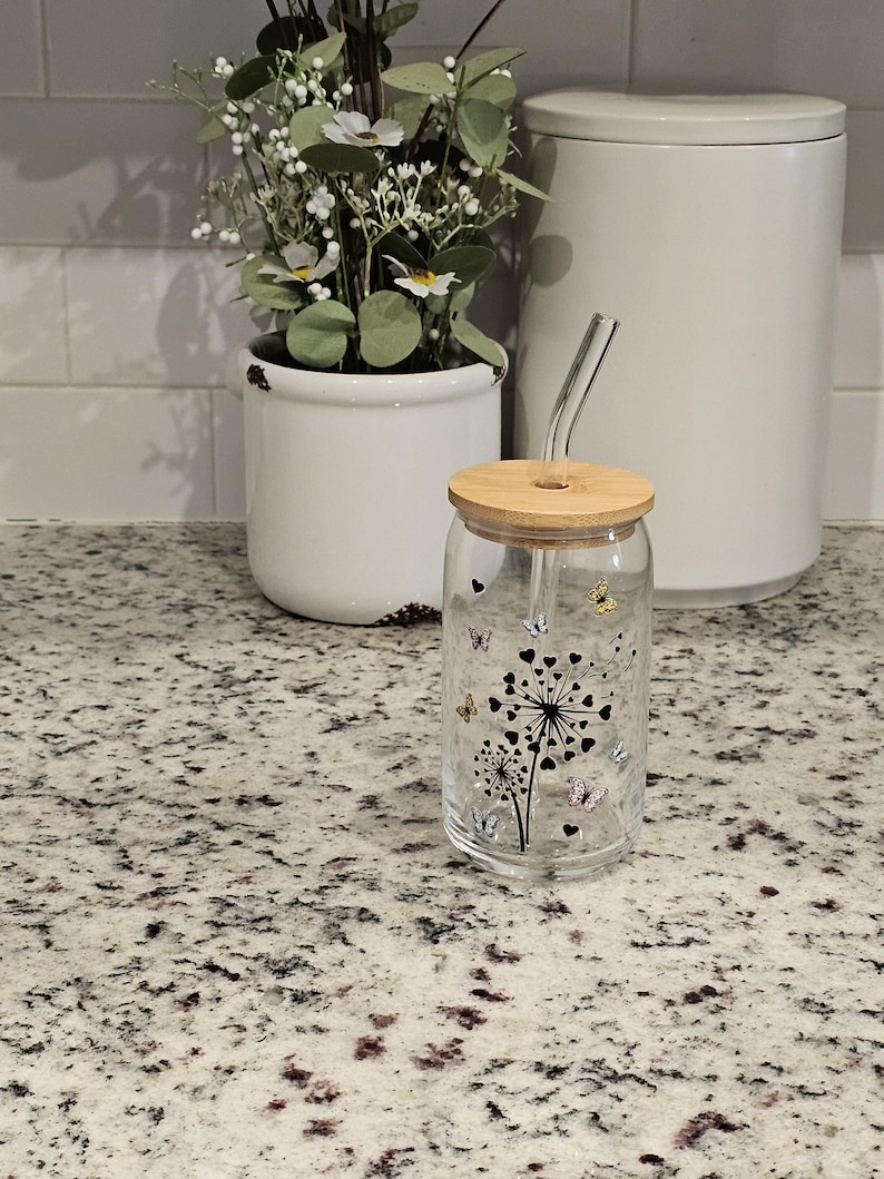 May include: A clear glass tumbler with a bamboo lid and a curved glass straw. The tumbler features a black dandelion design with colorful butterfly accents. A white ceramic pot with greenery and a white canister are in the background.