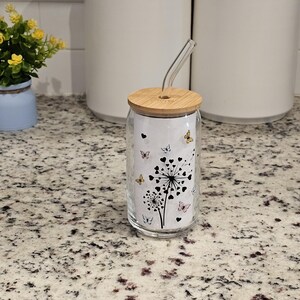 May include: A clear glass tumbler with a bamboo lid and a curved glass straw. The tumbler features a black and white dandelion design with butterflies in various colors. The tumbler is sitting on a speckled countertop.