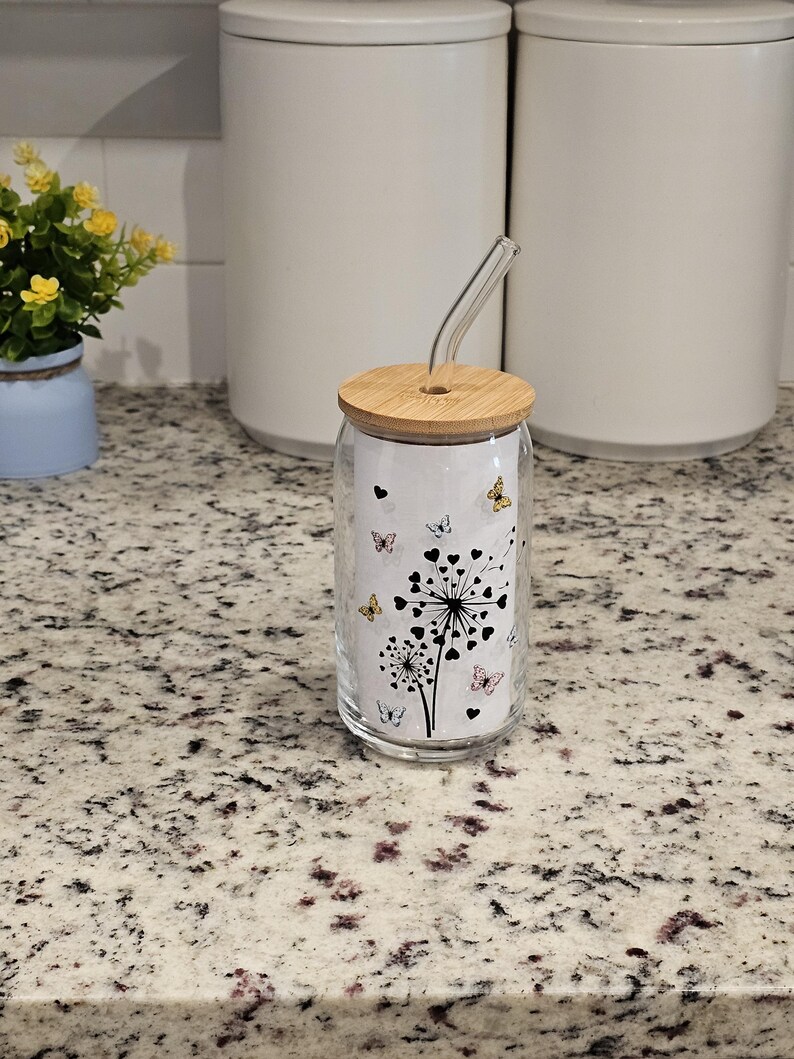 May include: A clear glass tumbler with a bamboo lid and a curved glass straw. The tumbler features a black and white dandelion design with butterflies in various colors. The tumbler is sitting on a speckled countertop.