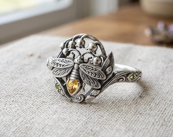 May include: A silver ring featuring a central bee motif with a pear-shaped yellow gemstone body. The design includes intricate wing detailing, small lily of the valley flowers, and tiny green and clear accent stones set along the band.