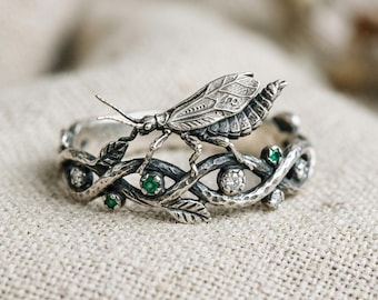 May include: A silver ring featuring a detailed insect perched on a band designed as intertwined branches. The band is decorated with small, round, clear white stones and vibrant green stones set within the textured, dark-toned metal branches.