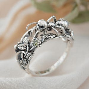 Könnte beinhalten: Ein silberner Ring mit einem Ameisen-Design, das auf einem Ast mit Blättern und Edelsteinen sitzt. Der Ring hat ein detailliertes, von der Natur inspiriertes Design mit poliertem Finish. Ein einzigartiges Schmuckstück.
