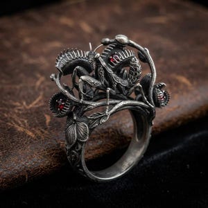 Op de afbeelding: Een zilveren ring met een gedetailleerde bidsprinkhaan die tussen Venusvliegenvangers zit. Het metaal heeft een geoxideerde afwerking met ingewikkelde texturen op de bladeren en het insectenlichaam. Kleine, ronde, dieprode edelstenen zijn gezet.