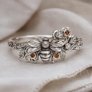 May include: A silver ring with a bee motif, showcasing intricate detailing on the insect's body and wings. The ring is adorned with floral elements, honeycombs, and small gemstones, set against a neutral backdrop.