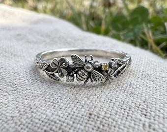 May include: A silver ring featuring a central bee motif surrounded by sculpted clover leaves and stems. The design includes a small yellow gemstone and two clear, colorless stones set among the metallic floral and insect details.