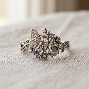 Könnte beinhalten: Ein silberner Ring mit einem detaillierten Design aus einem Schmetterling, einem Käfer und kleinen Blumen. Das Metall hat ein oxidiertes Finish mit kleinen, runden, violetten und klaren Edelsteinen, die in das florale Design eingearbeitet sind.