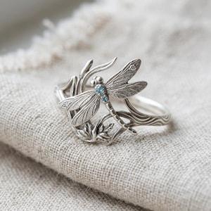 Puede incluir: Un anillo de plata con un diseño detallado de libélula, alas texturizadas y dos pequeñas piedras azul claro engastadas en el cuerpo. La banda del anillo incorpora líneas orgánicas y fluidas que se asemejan a tallos o hojas.