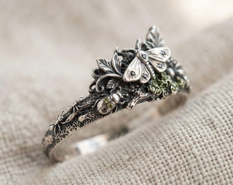 May include: A sterling silver ring featuring a detailed moth with black stone accents, a small beetle with green stone inlays, and textured leaf motifs. The band has a dark, oxidized finish with a rough, organic surface texture throughout.