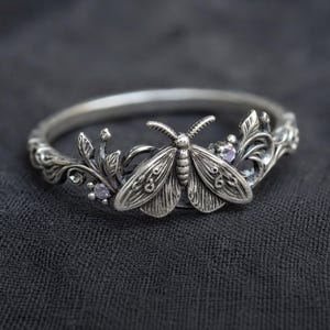 Puede incluir: Anillo de plata con un diseño detallado de polilla, con intrincados patrones de alas y hojas delicadas. Pequeñas gemas moradas están engastadas entre el follaje. La banda del anillo tiene un diseño ornamentado y texturizado.