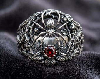 May include: A silver ring featuring a detailed spider design. The ring has a central spider figure, surrounded by an intricate floral and vine pattern. A round, red gemstone is set at the base of the spider. The ring is set against a dark background.