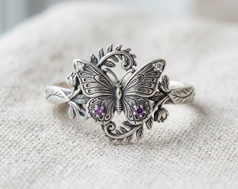 May include: A silver ring featuring a central butterfly motif with intricate wing detailing, accented by two small purple stones on the lower wings and two clear stones on the upper wings, surrounded by sculpted silver leaves and vine elements.