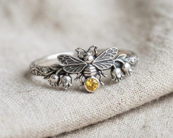 May include: A silver ring featuring a central bee motif with a round yellow gemstone set at the base of its abdomen. The band is decorated with sculpted floral elements resembling lily of the valley flowers extending from the bee's wings.