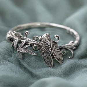 Könnte beinhalten: Silberfarbenes Armband mit detailliertem Zikaden-Design, einschließlich strukturierter Flügel und Kopf. Das Armband hat Blatt- und Rankenmotive sowie zwei kleine grüne Edelsteine. Der Schmuck liegt auf einem teal-farbenen Stoff.
