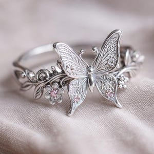 Puede incluir: Anillo plateado con diseño de mariposa. El anillo presenta una mariposa detallada con detalles brillantes y pequeñas gemas rosas. Los diseños florales y de hojas adornan la banda, creando una pieza de joyería elegante e inspirada en la naturaleza.