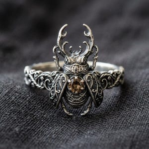 Puede incluir: Anillo plateado con un diseño detallado de escarabajo. El escarabajo tiene intrincados patrones en alas y cuerpo, con una gema marrón central. La banda del anillo presenta un patrón decorativo y ondulado.