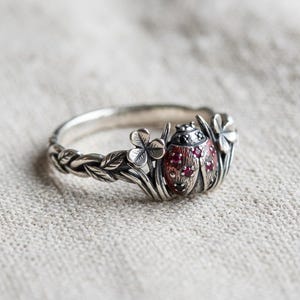 Peut inclure: Une bague en argent ornée d'un motif de coccinelle avec de petites pierres rouges sur les ailes et des détails en pierres noires sur la tête. L'anneau présente un motif de vigne et de feuilles, entouré de petits trèfles à trois feuilles.