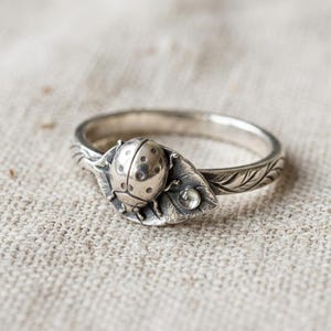 Può includere: Un anello in argento con una coccinella scolpita appoggiata su una foglia testurizzata. La fascia è dettagliata con venature di foglia incise e una piccola pietra preziosa rotonda e trasparente è incastonata sulla foglia accanto alla coccinella.