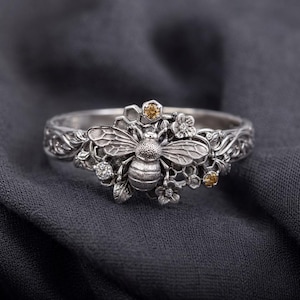 May include: A silver ring with a bee motif, honeycomb, and floral details. The ring includes small gemstones and is set against a dark grey backdrop. The band of the ring has an intricate, engraved pattern.