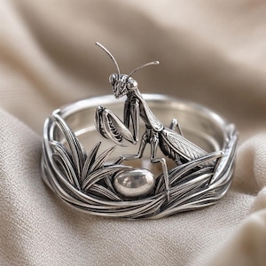 May include: A silver ring featuring a detailed praying mantis perched atop a band sculpted to resemble blades of grass, with a small, smooth, polished silver sphere nestled among the textured foliage at the base of the insect.