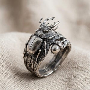 May include: A silver ring featuring a detailed stag beetle perched on a textured branch design. The band incorporates a small acorn element, with an oxidized finish that highlights the intricate carvings of the insect and the wood grain texture.