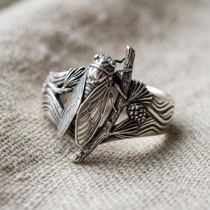 May include: A silver ring featuring a detailed cicada insect resting on a textured branch. The band incorporates relief carvings of pine needles and a small pinecone, with oxidized detailing that highlights the insect's wings and body structure.