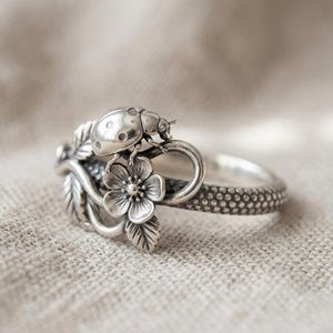 Könnte beinhalten: Ein silberner Ring mit einem Marienkäfer und floralen Motiven. Der Ring zeigt einen detaillierten Marienkäfer, eine Blume und Blätter. Das Band hat ein strukturiertes Muster. Ein Schmuckstück zum Thema Natur.