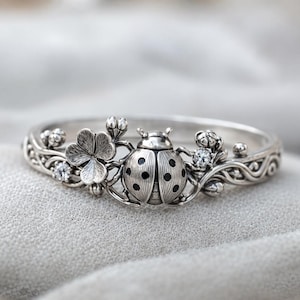 May include: A silver ring featuring a detailed ladybug design with black spots, a four-leaf clover, and small floral accents. The ring has a delicate, nature-inspired aesthetic, perfect for everyday wear.