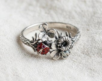 May include: A sterling silver ring featuring a textured band with a nature-inspired design. The ring displays a red ladybug with a small pink stone, a silver caterpillar, and several clover leaves. The interior of the band is stamped with 925.