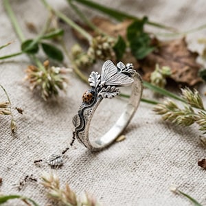 Può includere: Un anello in argento con fascia testurizzata, decorato con una piccola coccinella dettagliata a pois neri e una falena con ali incise. L'anello presenta motivi a foglia e un delicato ciondolo pendente a forma di foglia fissato a una sottile catenina.