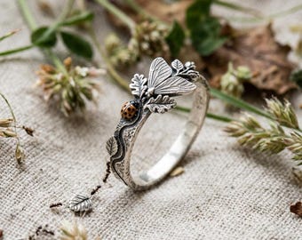 May include: A silver ring featuring a textured band with a small, detailed ladybug with black spots and a moth with etched wings. The ring includes small leaf motifs and a delicate dangling charm shaped like a leaf, attached by a thin chain.